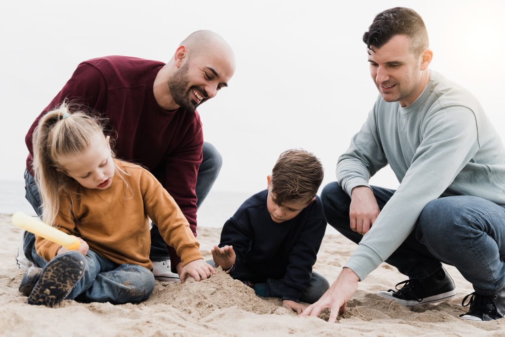 Gay male parents having fun with their children outdoor at the beach - Lgbt family playing together